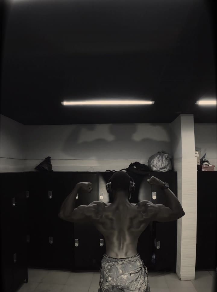 Coach TYGR from behind in a dark locker room, holding a back double-biceps pose.